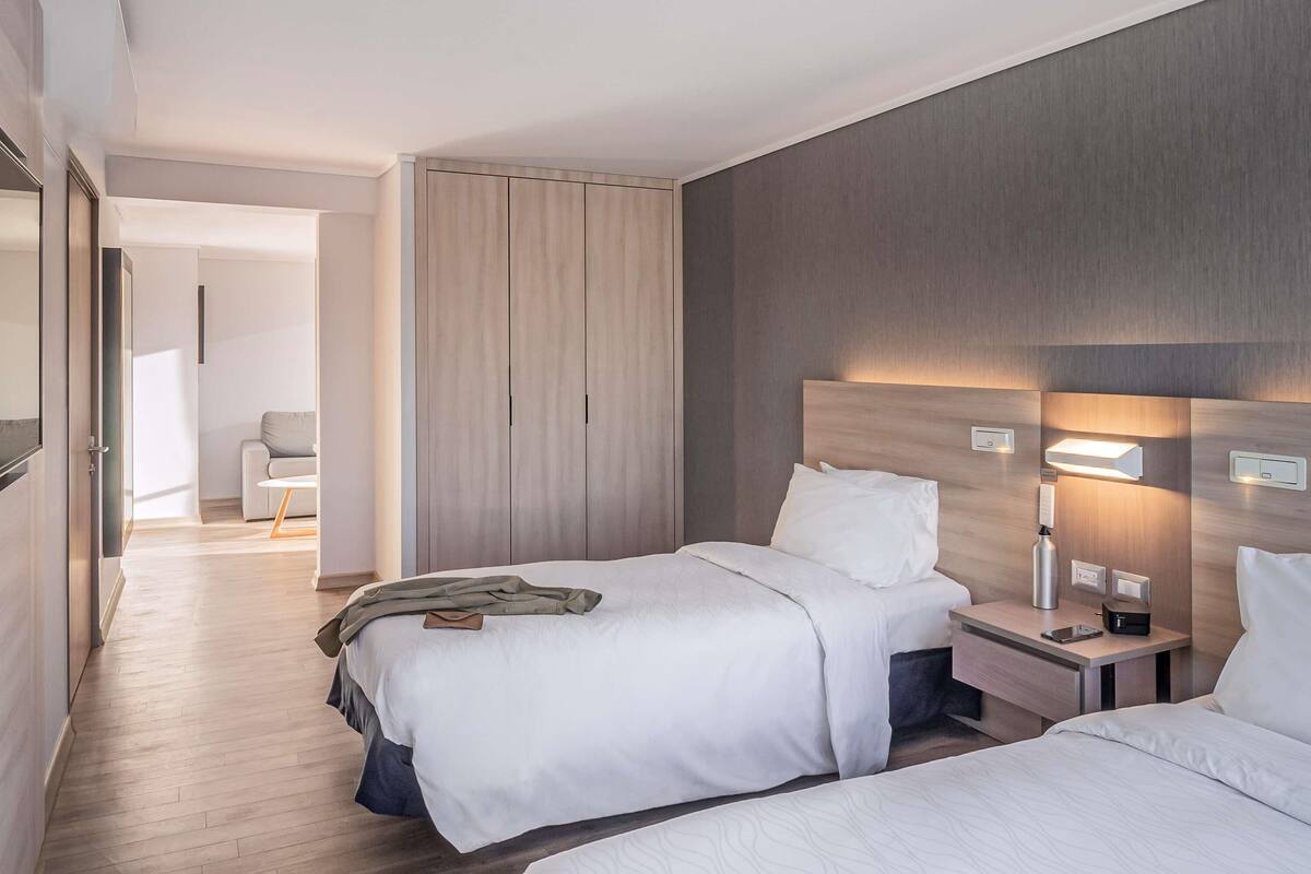 A hotel room with two beds, a wardrobe, a bedside table with a lamp, and a view of the hallway.