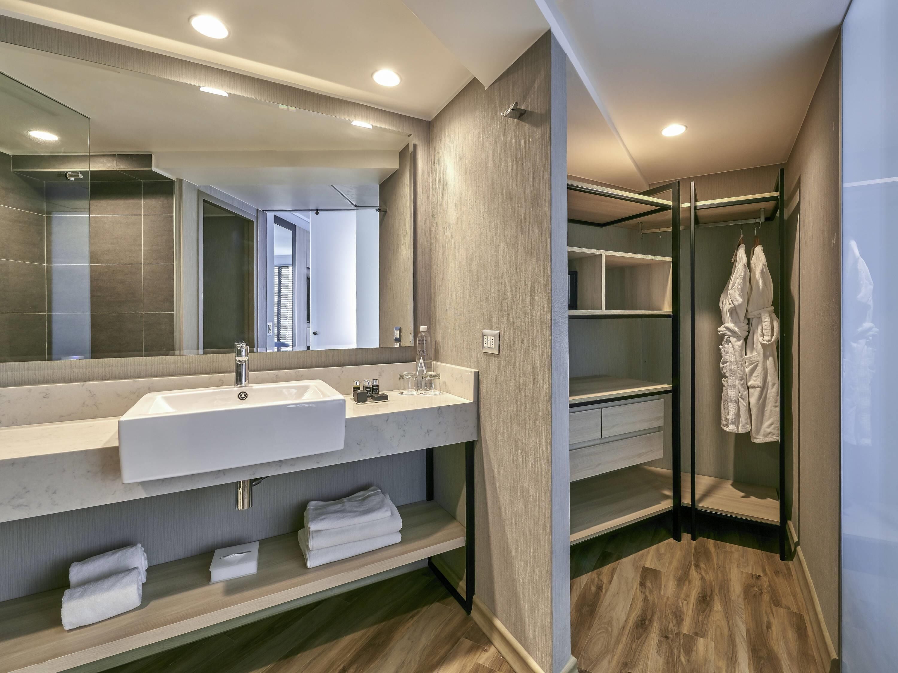 A modern bathroom with a large mirror, two sinks, a shower, and a walk-in closet with shelves.