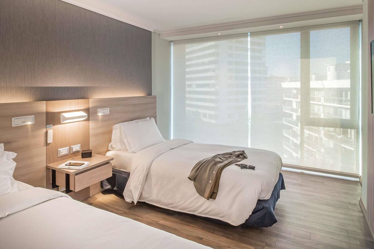 A modern hotel room with a large window, a bed, a bedside table, and a nightstand.