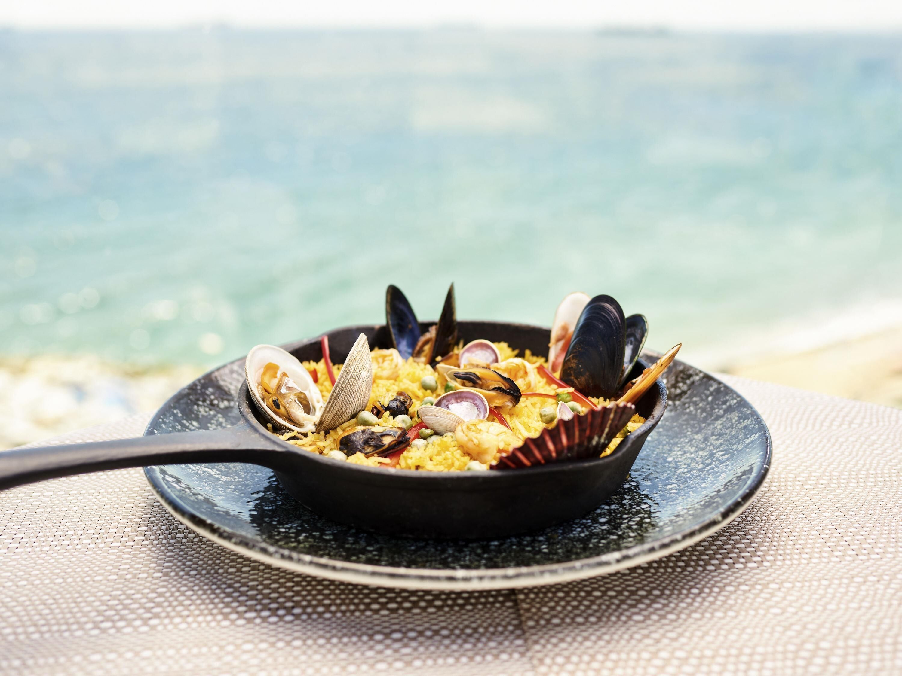 A seafood dish in a black skillet on a table with a textured surface, set against a blurred background of a body of water.