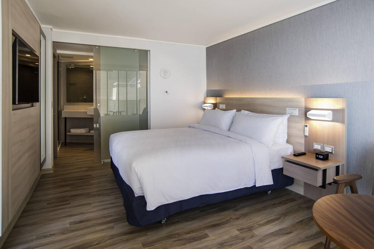 A modern hotel room with a bed, bedside tables, a TV, and a bathroom visible through a glass door.
