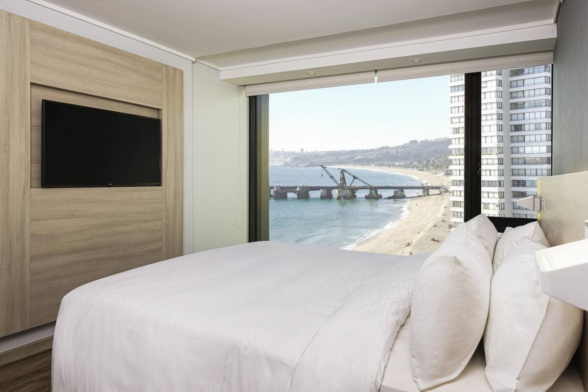 A modern hotel room with a large bed, a TV mounted on the wall, and a view of a beach and bridge.