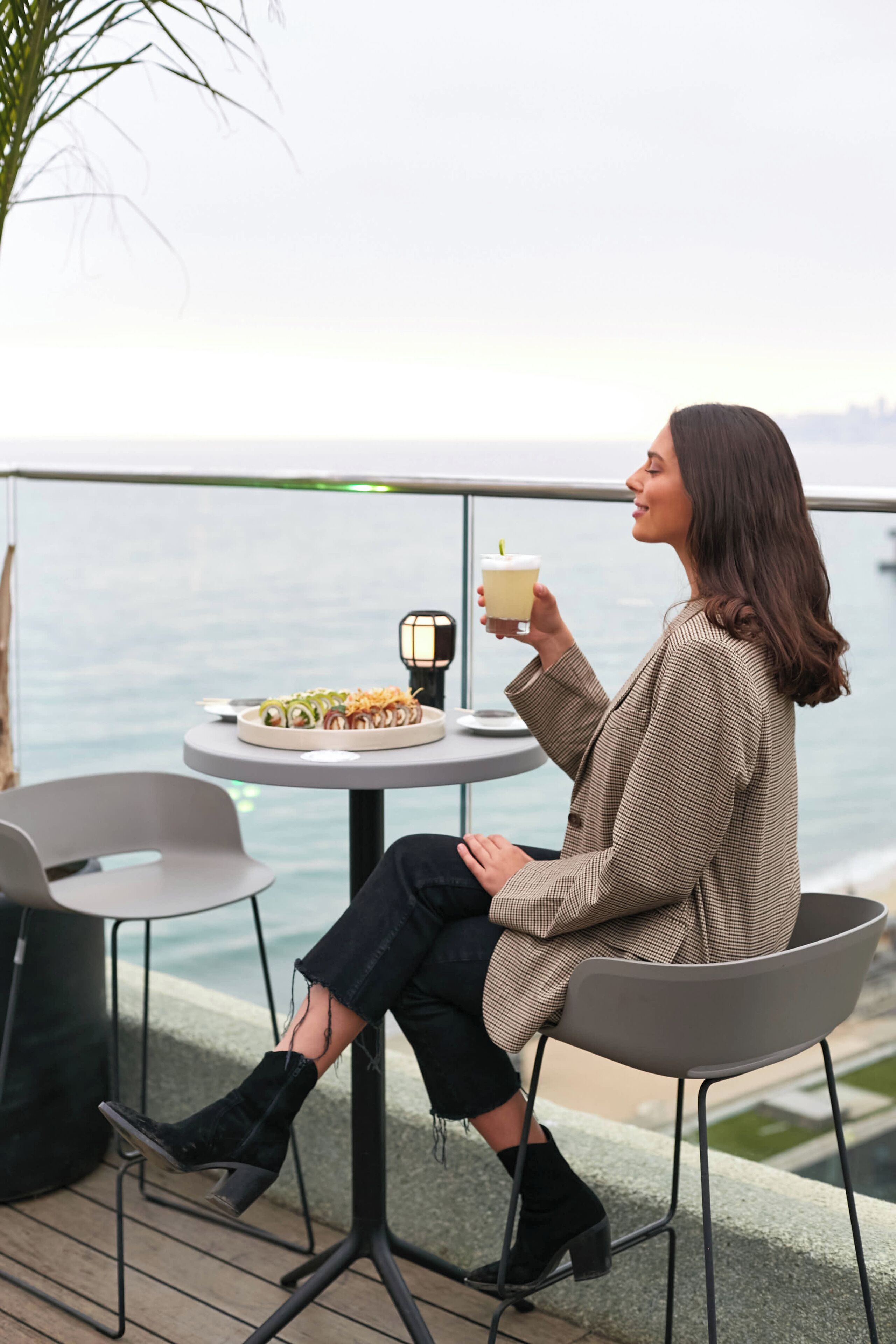 A person seated at a round table with a plate of food and a drink, overlooking a body of water.