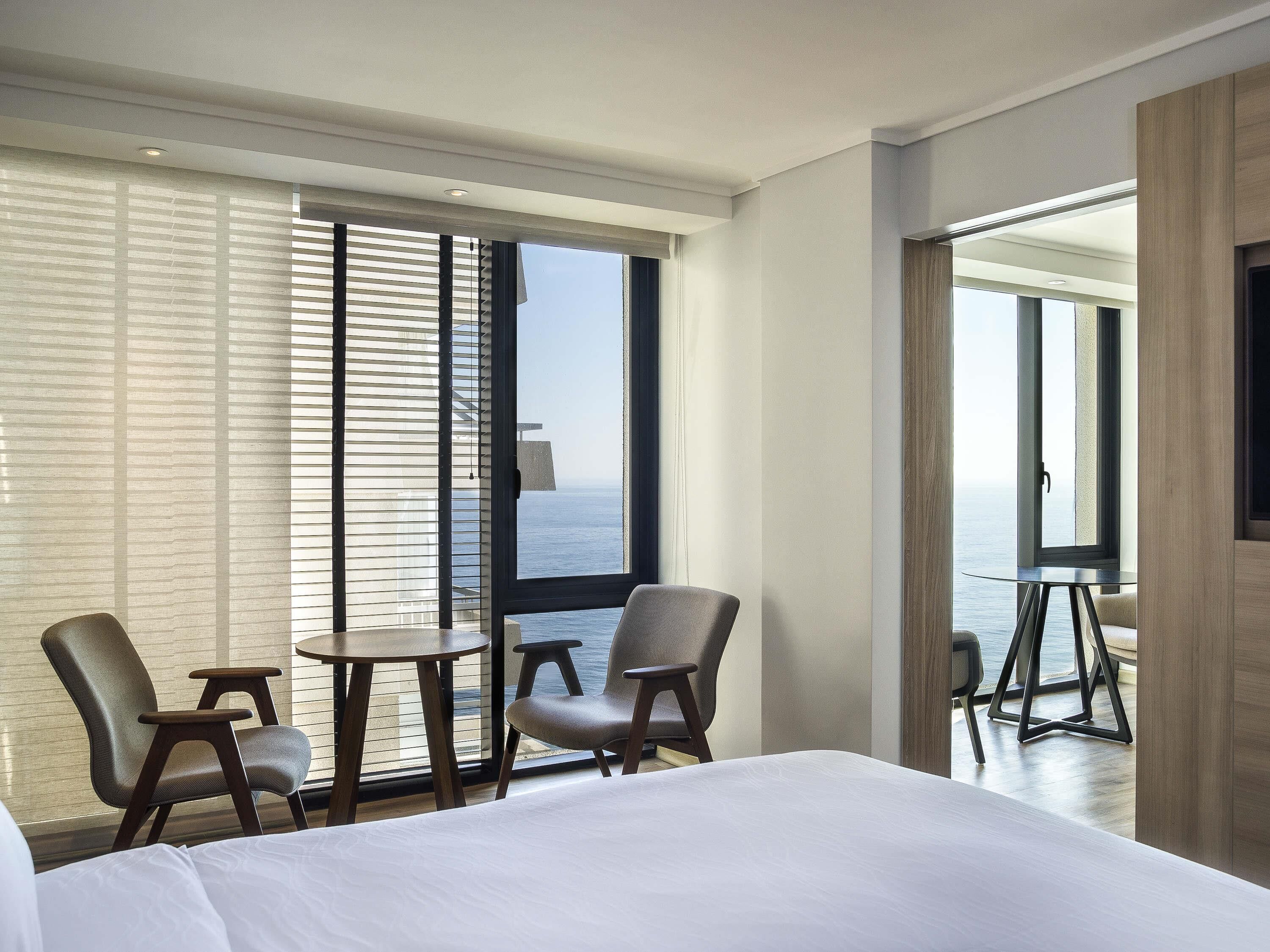 A hotel room with a bed, two chairs, a small table, and a view of the sea through large windows.