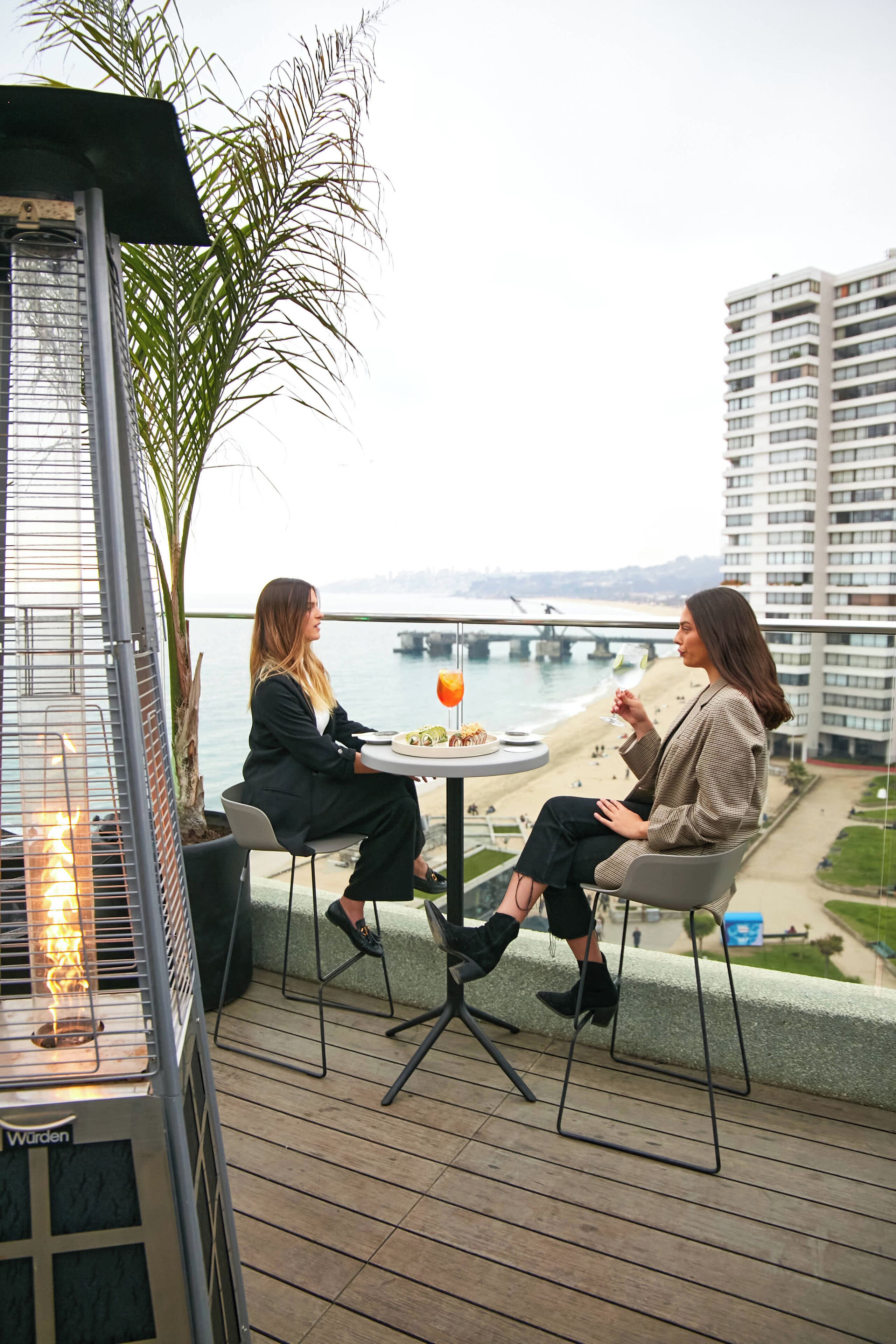 Dos personas sentadas en una mesa en un balcón con vista a una playa y un puente.