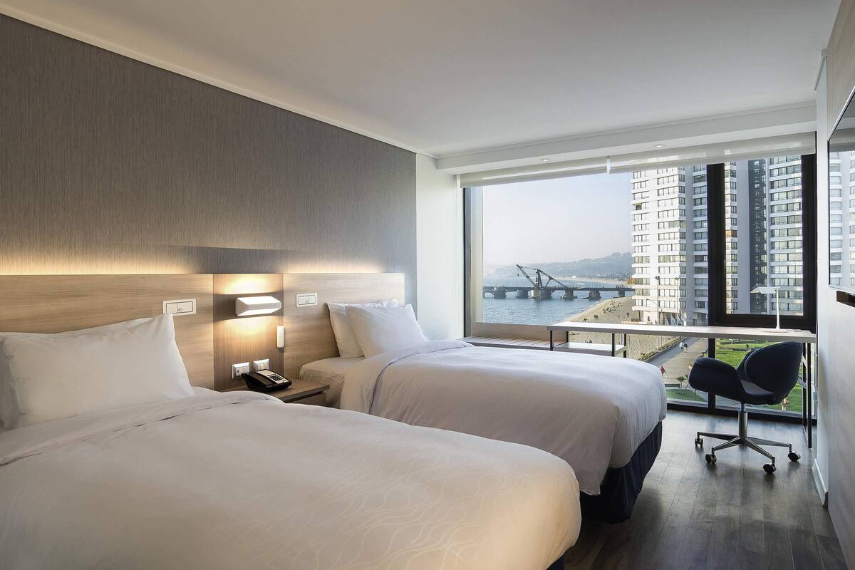A hotel room with two beds, a nightstand, a desk, and a large window with a view of a bridge and buildings.