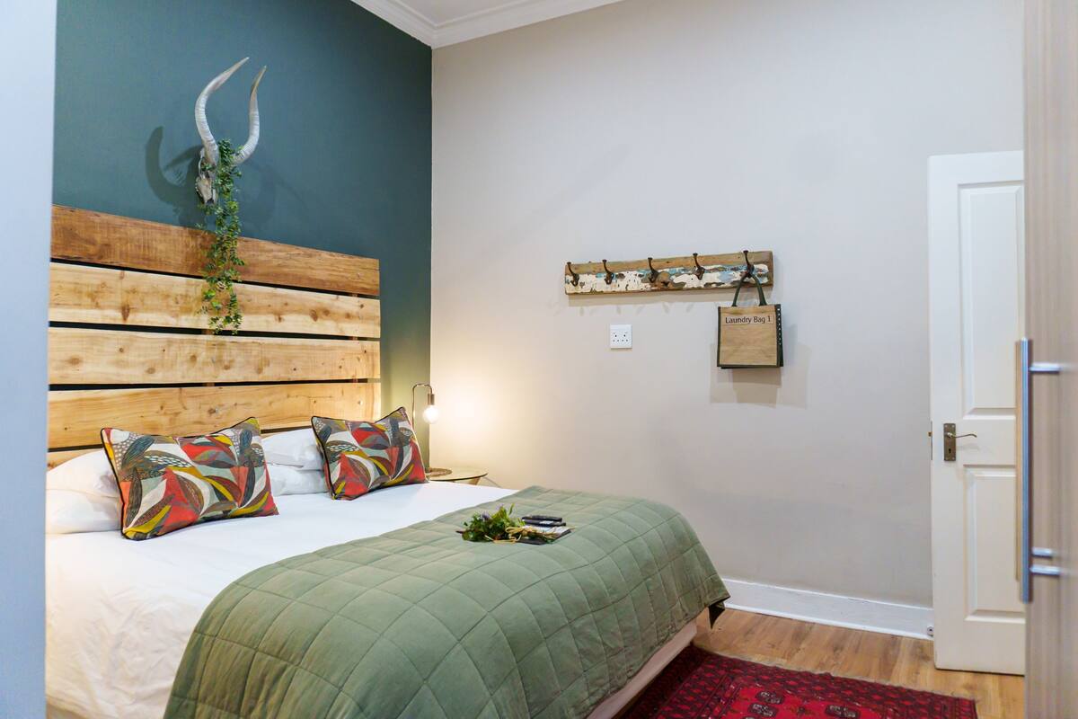 A bedroom with a wooden headboard, a bed with a green quilt, decorative pillows, a wall-mounted coat rack, and a door.