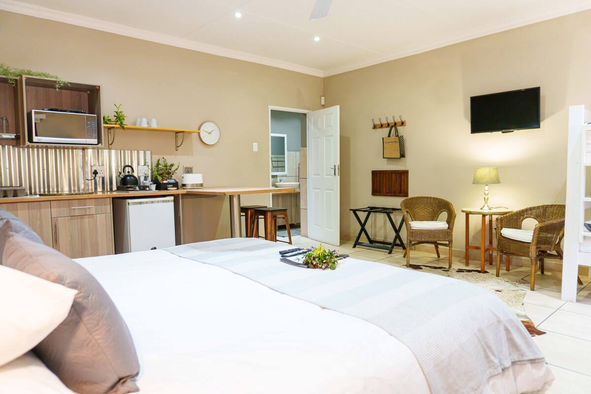 A hotel room with a bed, a kitchenette, a dining area with chairs, a TV, and a clock on the wall.
