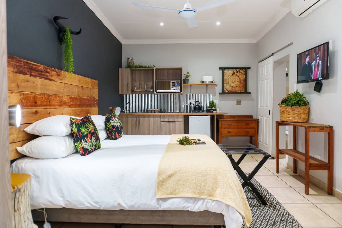 A bedroom with a bed, pillows, a bedside table, a wooden headboard, a ceiling fan, a television, and a kitchen area with a microwave, refrigerator, and cabinets.