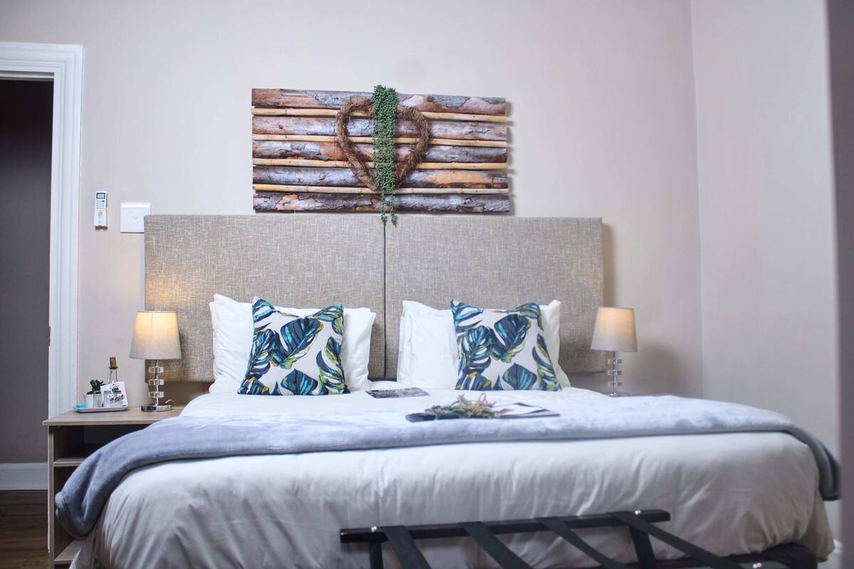 A hotel room with a large bed, two bedside lamps, a nightstand with a lamp, a decorative wall piece, and a door.
