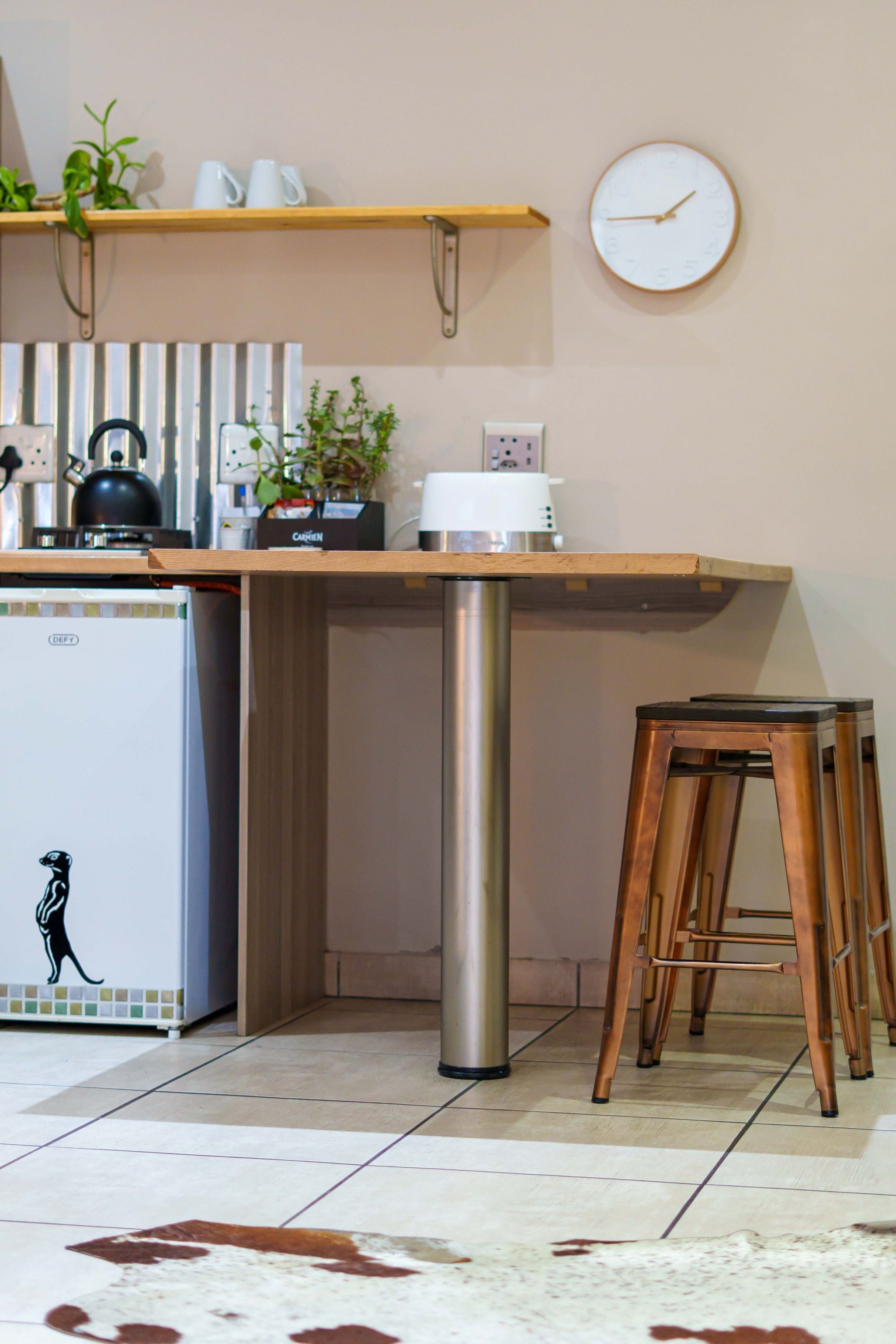 Een keuken met een koelkast, waterkoker en koffiezetapparaat, een houten kruk en een klok aan de muur.