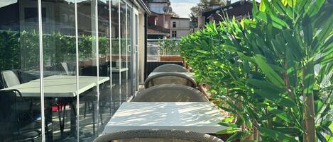A sunlit outdoor dining area with a glass wall, wicker chairs, and a wooden planter with green plants.