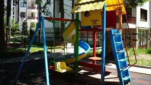 A playground with a slide and climbing structures, surrounded by trees and buildings. - Apart Hotel Flora Residence (Samokov)