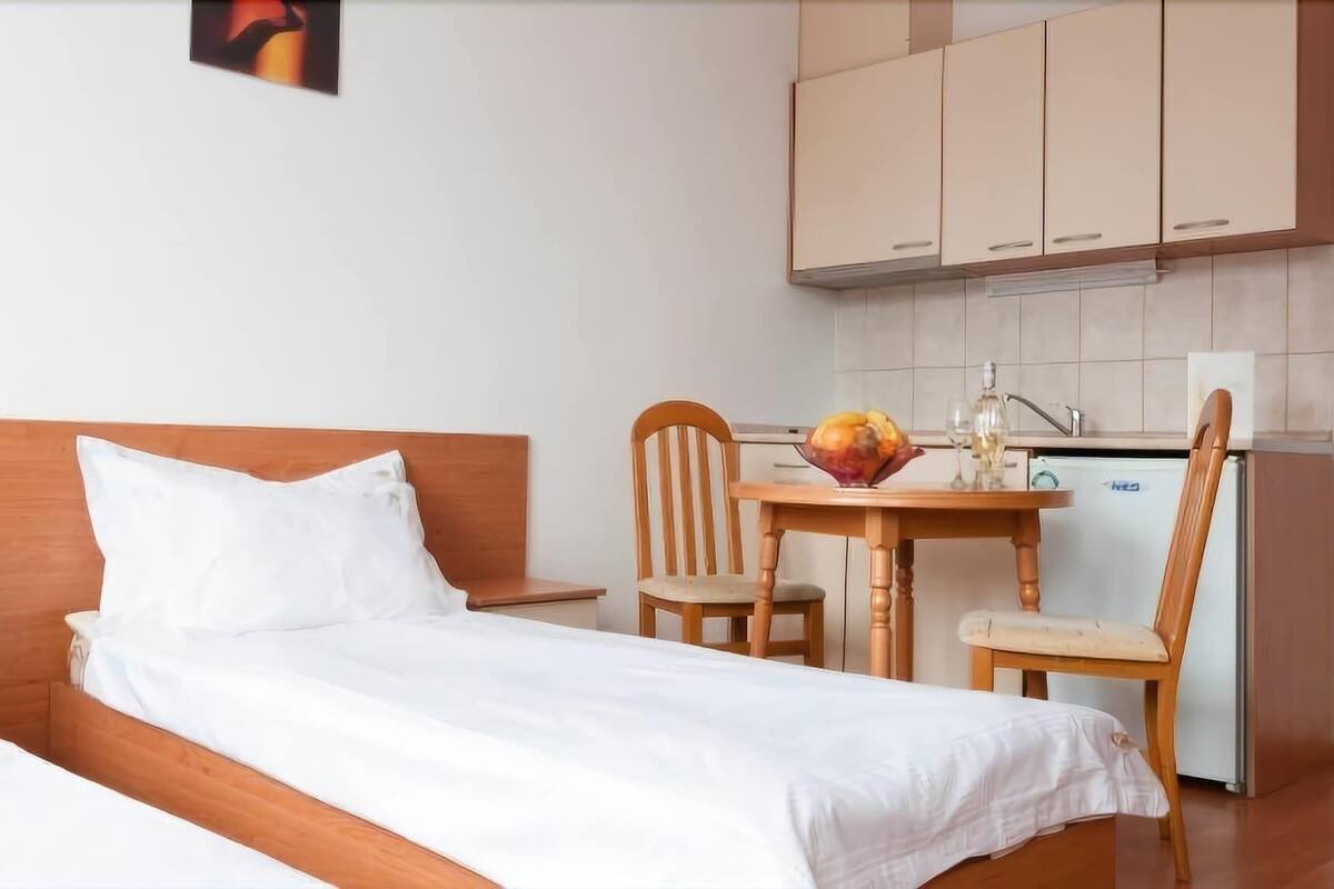 Una habitación pequeña y limpia con una cama, una mesa de madera con sillas, una zona de cocina con armarios y un refrigerador.