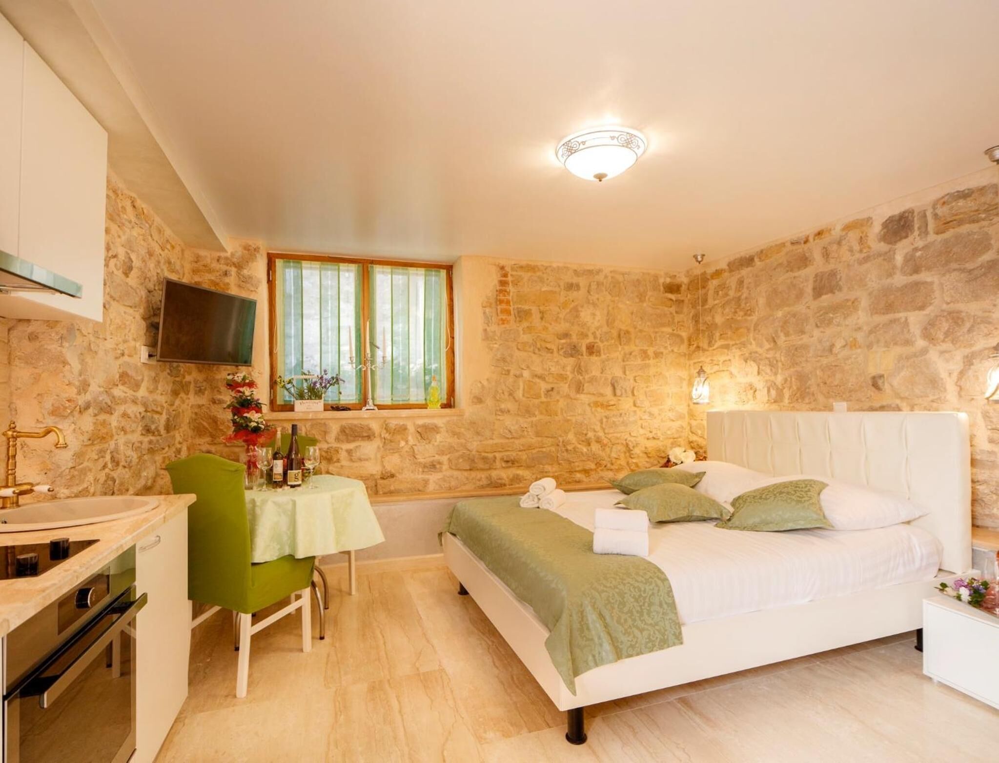 A bedroom with a stone wall, a bed with green bedding, a bedside table, a small table with a chair, and a TV mounted on the wall.