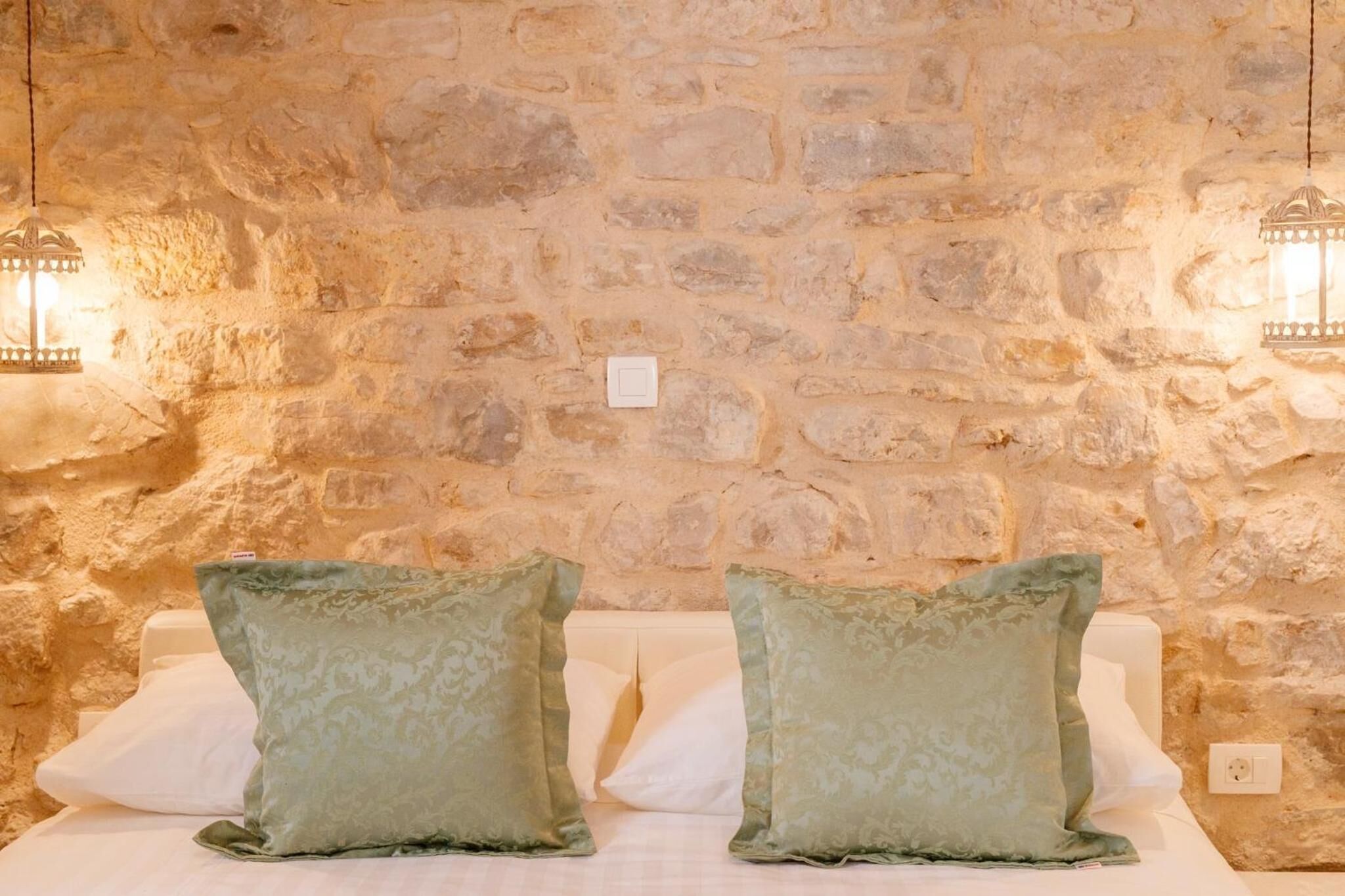 A room with a stone wall, two wall-mounted lights, and two green pillows on a white bed.