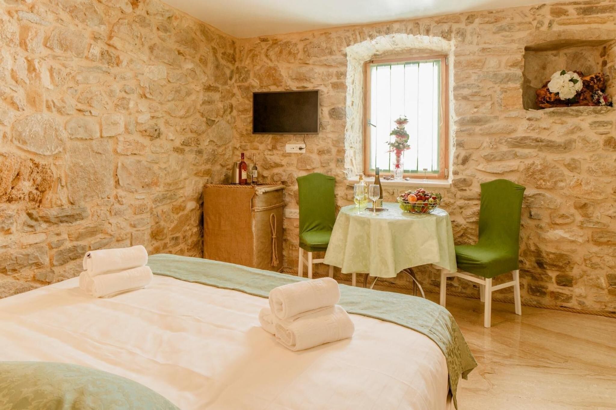 Quarto com paredes de pedra, uma cama, duas cadeiras verdes, uma mesa pequena com toalha de mesa verde, uma televisão fixada na parede e uma janela com vista para flores.