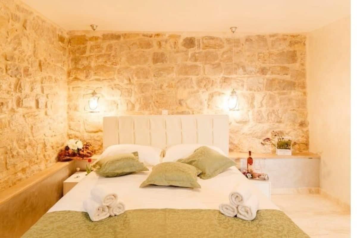 A bedroom with a stone wall, a bed with green pillows, a bedside table with a lamp, and a small plant.