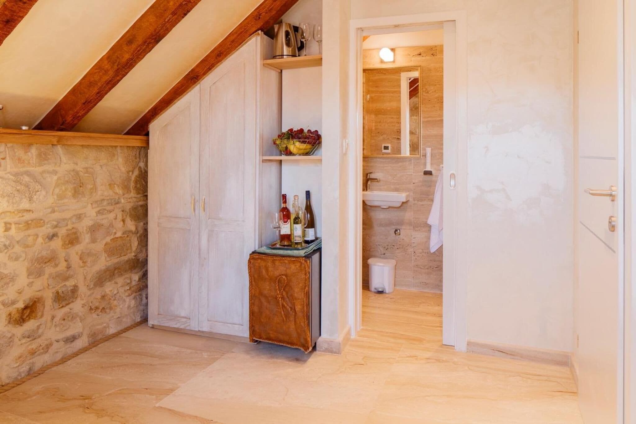 A room with a stone wall, wooden beams, a white wardrobe, a small wooden cabinet with bottles, and a bathroom with a sink and mirror.
