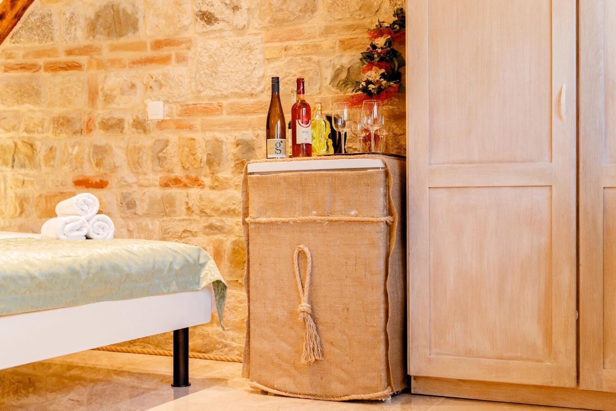 A room with a stone wall, a bed with a green and white bedspread, a wooden cabinet, and a small wooden chest with bottles and glasses on top.