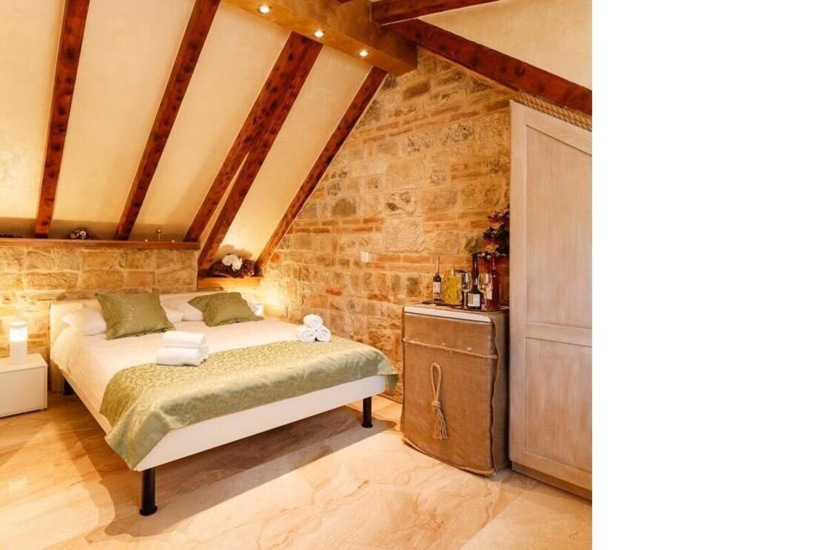 A bedroom with a stone wall, wooden beams, a bed with green bedding, a nightstand, and a cabinet.