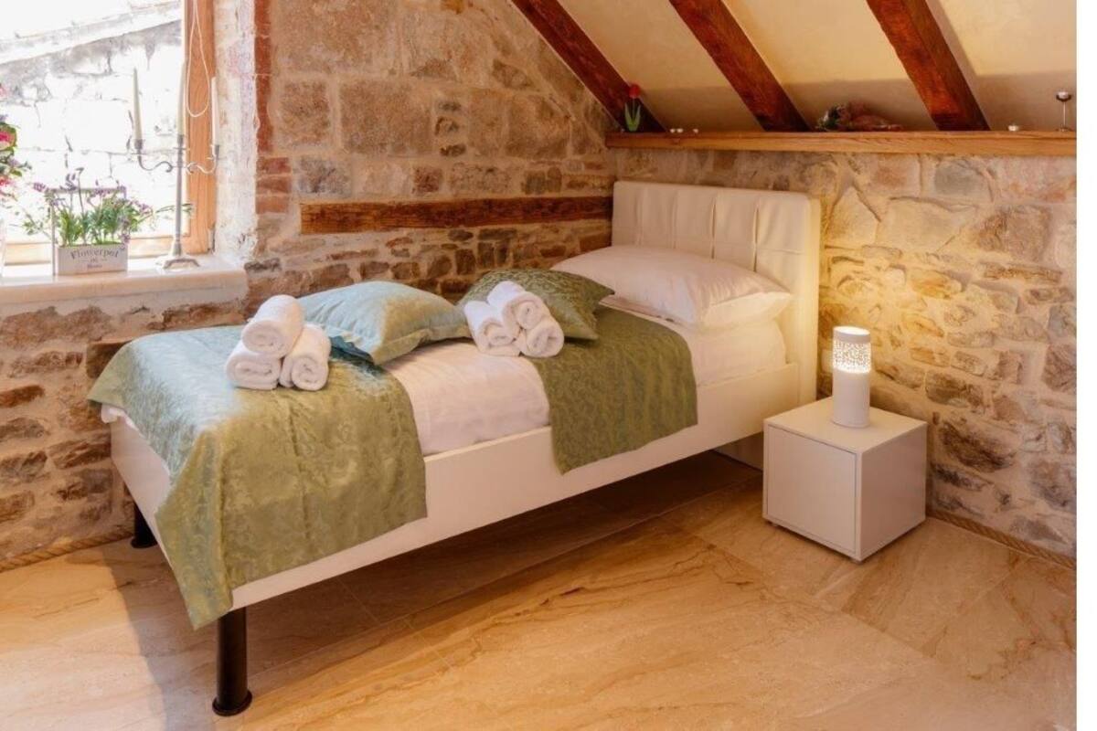 A cozy bedroom with a stone wall, a wooden ceiling beam, a bed with green bedding, and a bedside table with a lamp.