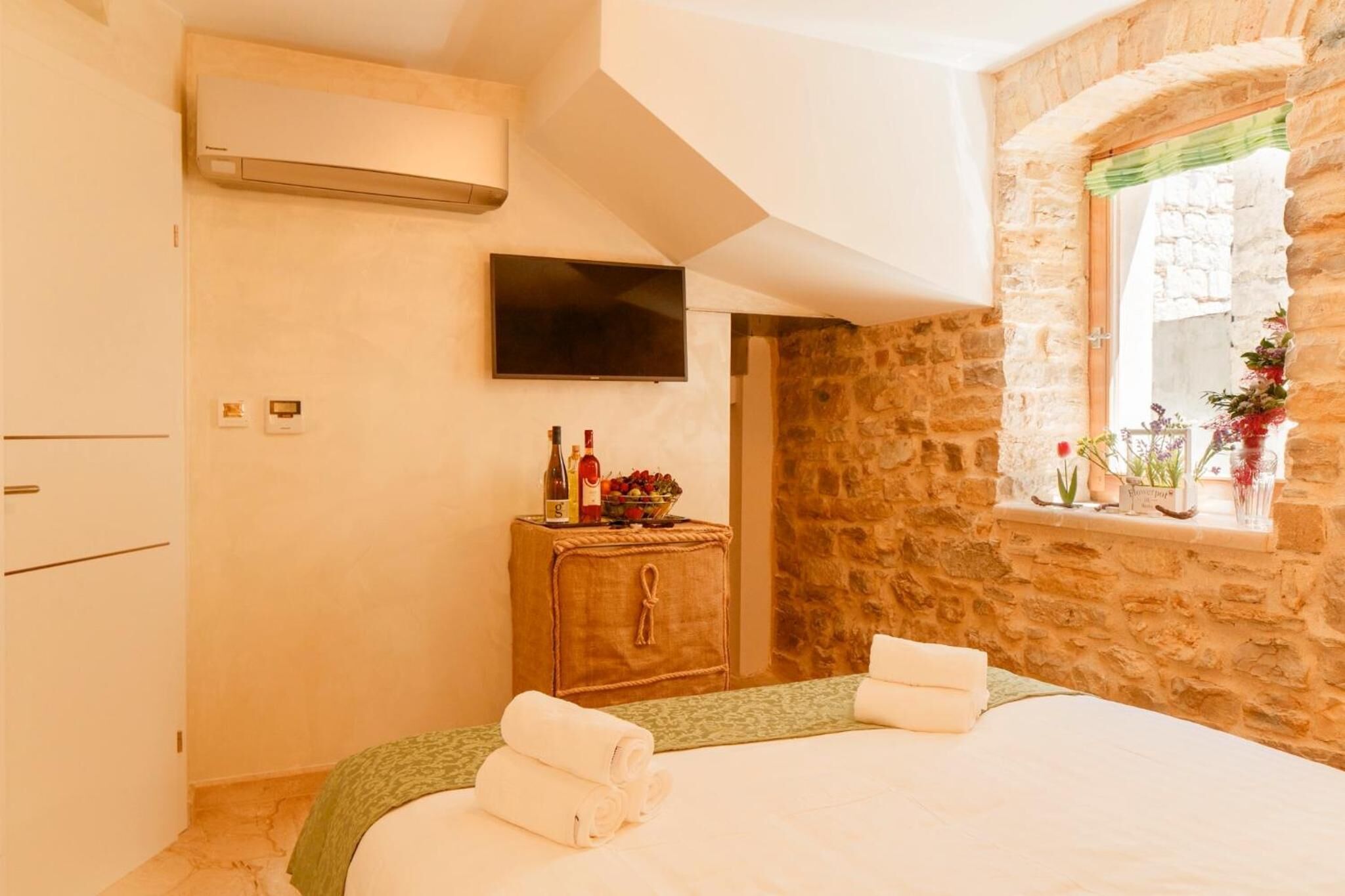 Un dormitorio con pared de piedra, un mueble de madera con botellas y fruta, un televisor de pantalla plana y una ventana con flores.