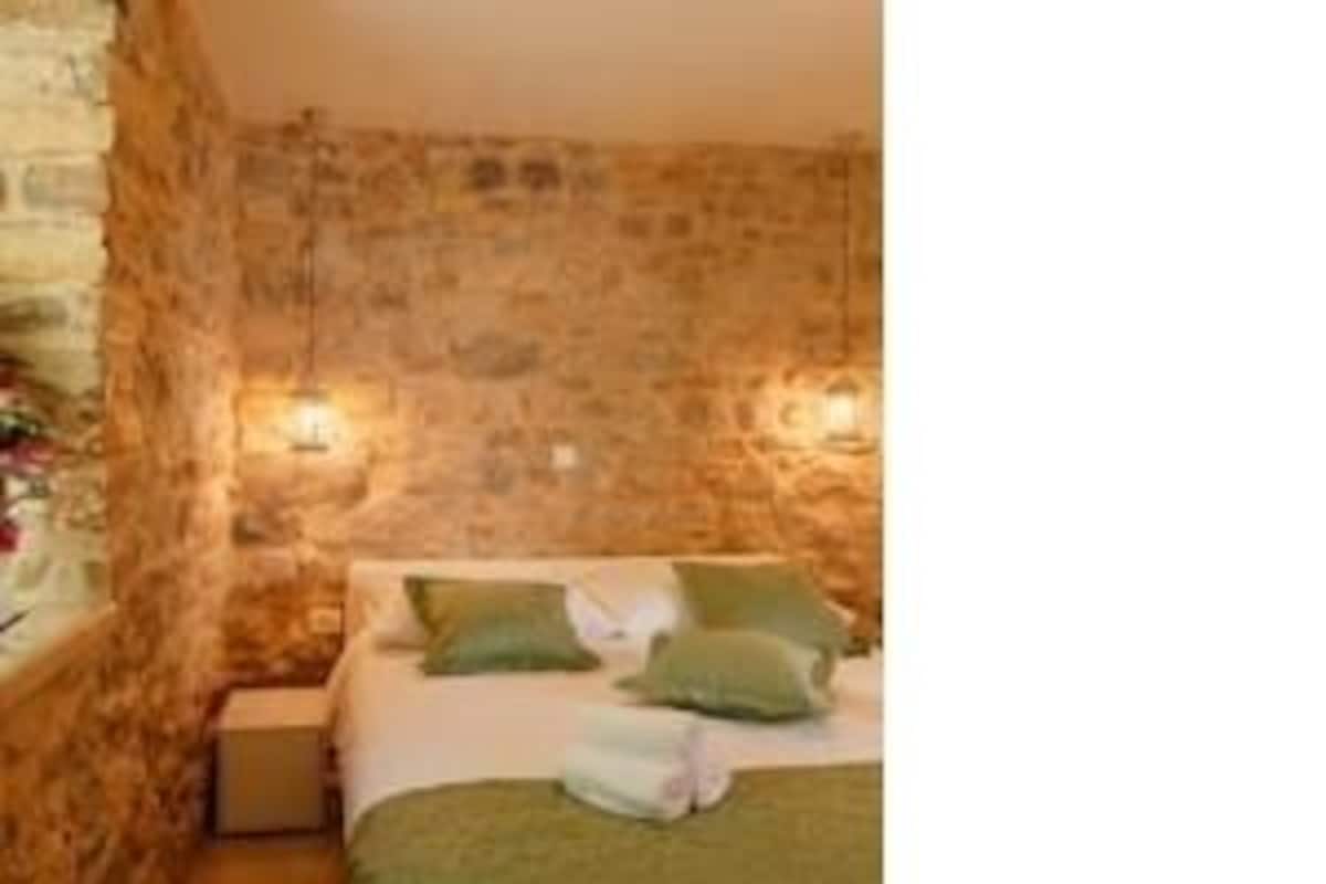 A room with a stone wall, a bed with green bedding, and a nightstand with a lamp.