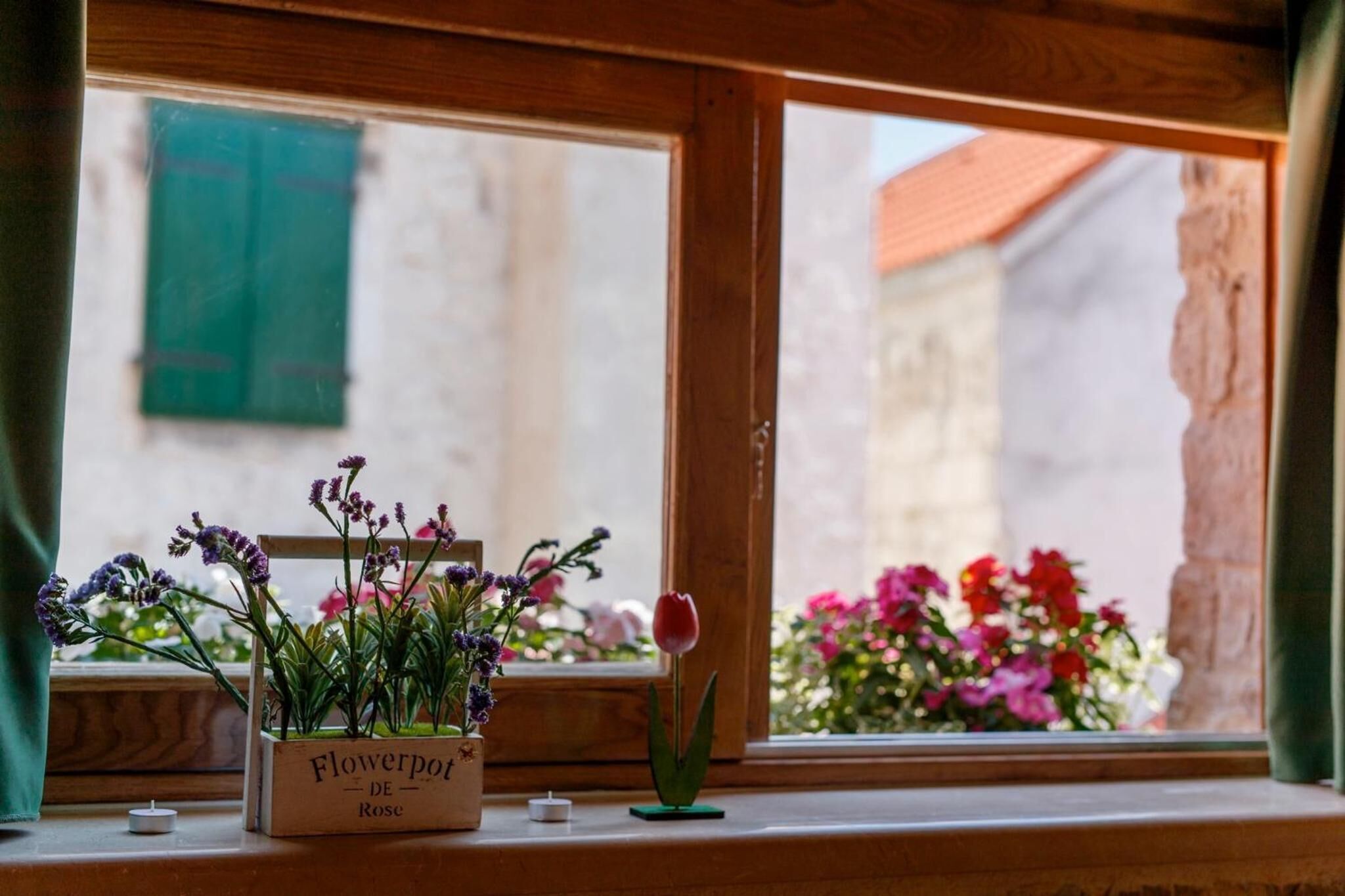 Una ventana con marco de madera, una pequeña caja de madera con la etiqueta "Flowerpot DE Rose" y una sola tulipa roja.