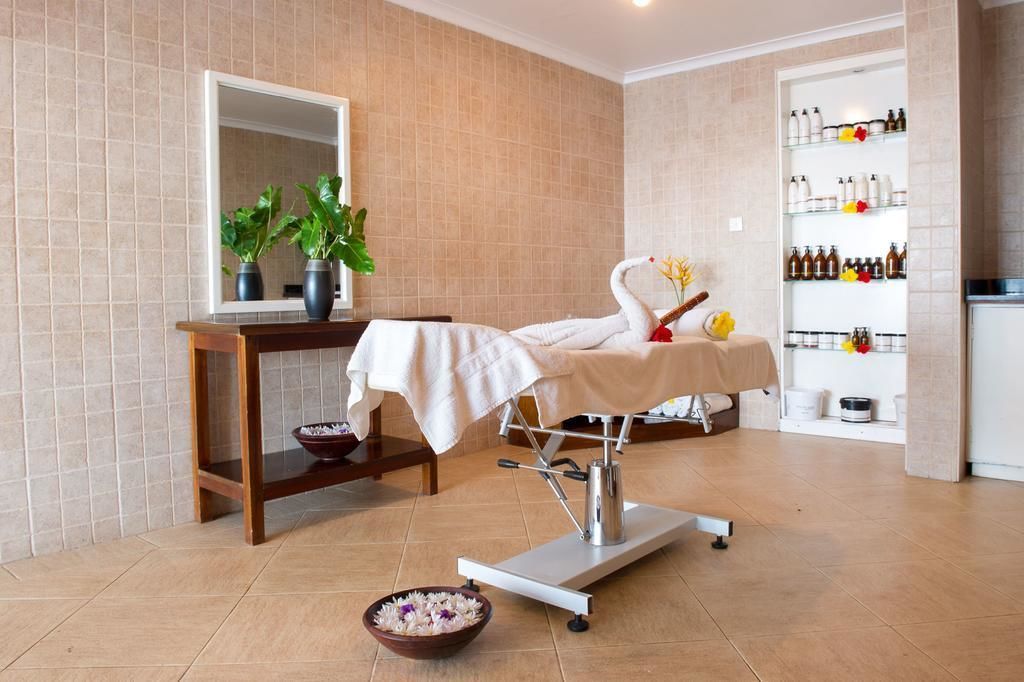 A spa room with a massage table, a wooden side table with a bowl, a mirror, and a shelf with bottles.