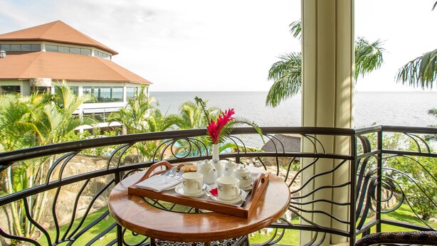 A balcony with a tray of white cups and saucers, a vase with red flowers, and a view of the ocean and a building with a red roof. - Malaika Beach Resort (Mwanza)