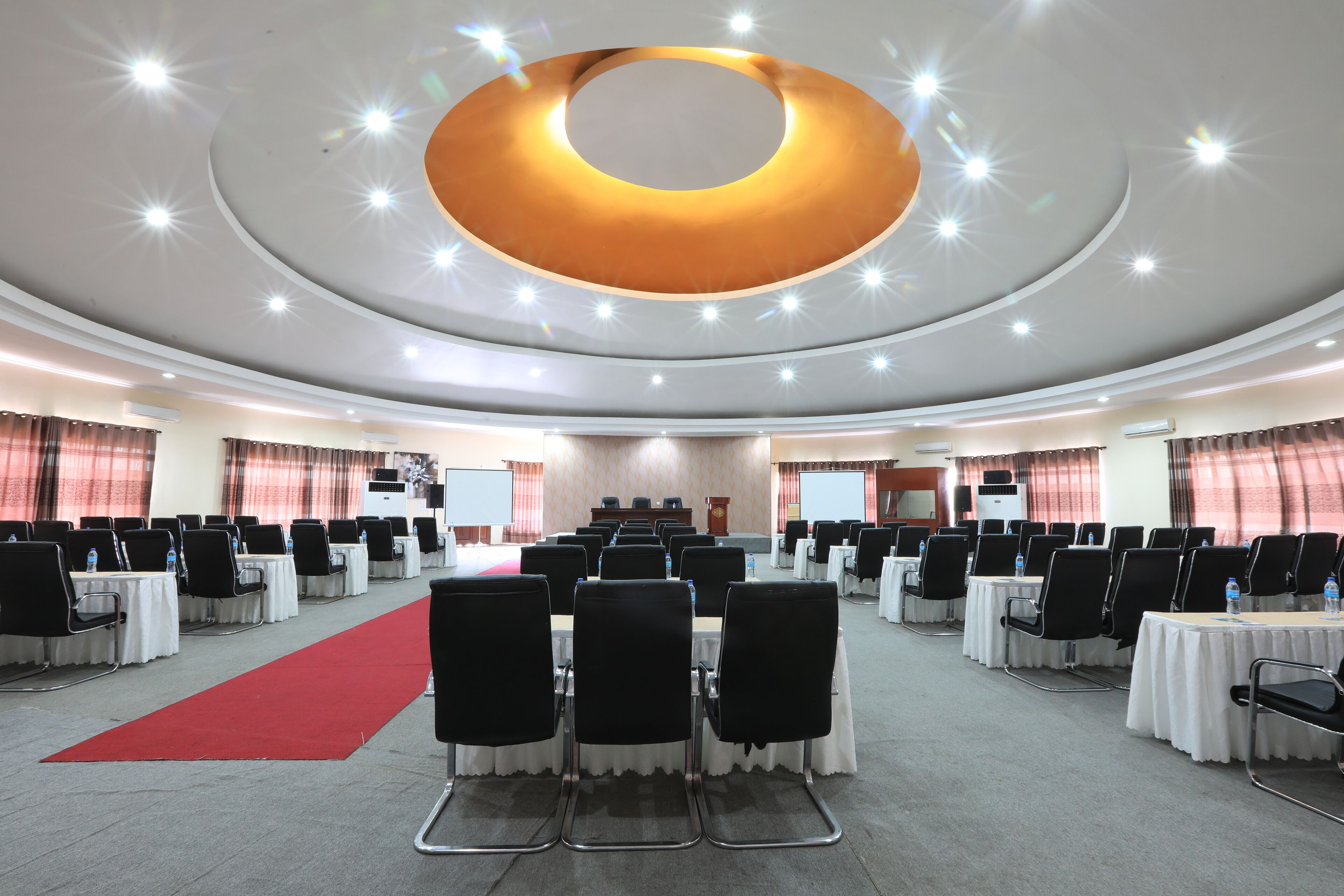A conference room with rows of tables and chairs, a red carpet, and a circular light fixture on the ceiling.