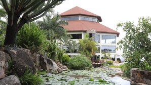 A building with a red roof and multiple windows, surrounded by lush greenery and a pond with lily pads. - Malaika Beach Resort (Mwanza)