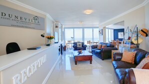 A reception area with a white reception desk, a sign reading "DUNEVILLA LUXURY GUEST HOUSE & BEAUTY SALON," and a view of the ocean through large windows. - Dune Villa Wilderness (Wilderness)