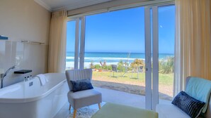 A bathroom with a bathtub, a chair, a sofa, and a view of the beach through large glass doors. - Dune Villa Wilderness (Wilderness)
