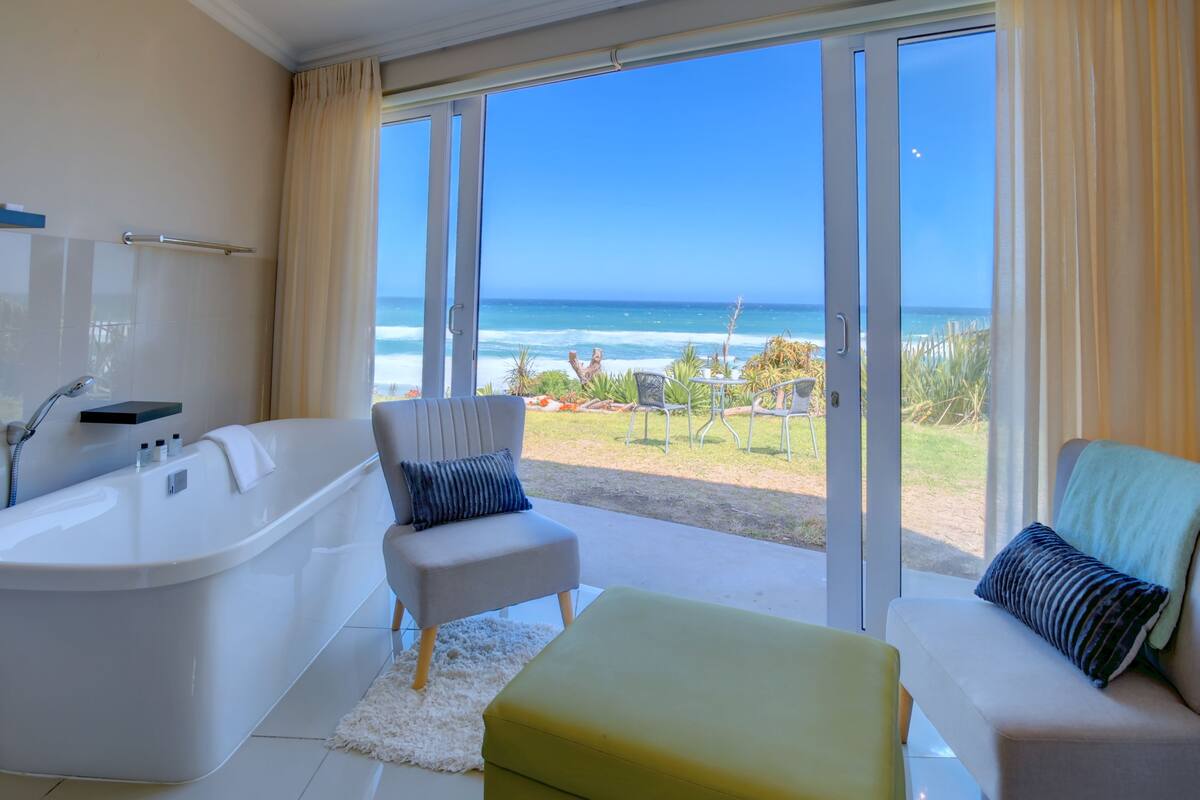 A bathroom with a bathtub, a chair, a sofa, and a view of the beach through large glass doors.