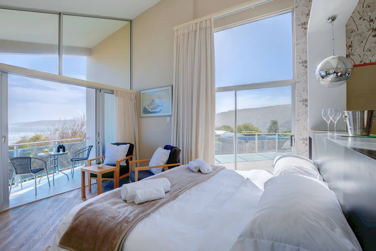 A hotel room with a large bed, a small table with chairs, a balcony with a table and chairs, and a view of the landscape.