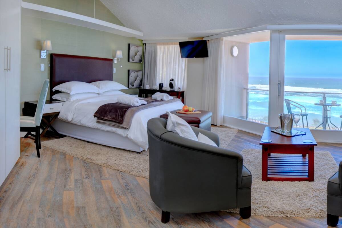 A modern hotel room with a large bed, two armchairs, a small table, and a balcony with a view of the ocean.