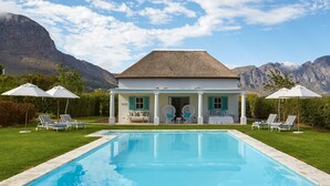 A poolside view of a house with a pool, lounge chairs, and umbrellas, surrounded by mountains. - La Cotte House (Franschhoek)