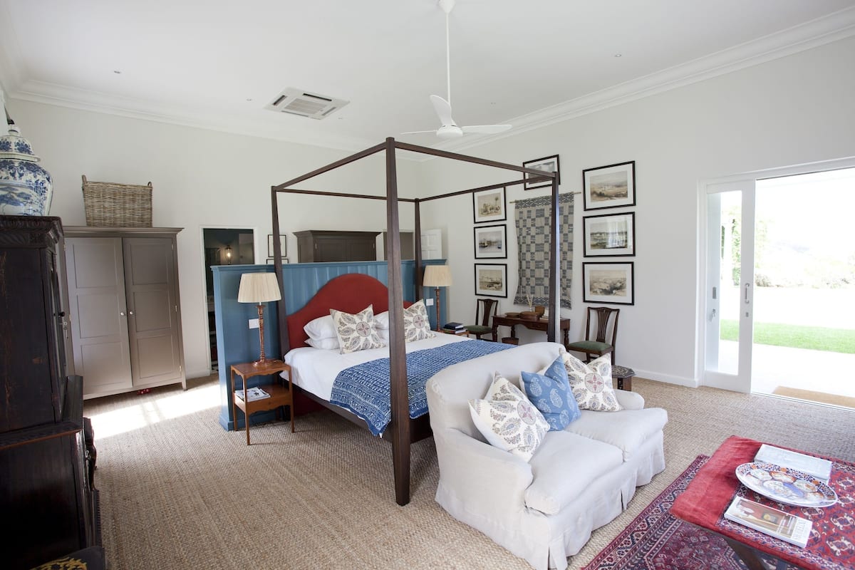 Un dormitorio con cama con dosel, una zona de estar con sofá blanco y vistas al exterior a través de un gran ventanal.
