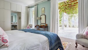 A bedroom with a bed, a desk, and a chair. There is a window with curtains and a view of greenery outside. - La Cotte House (Franschhoek)