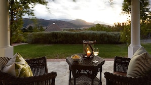 An outdoor seating area with wicker furniture, a candle, and a glass of wine, overlooking a scenic landscape with hills and a clear sky. - La Cotte House (Franschhoek)