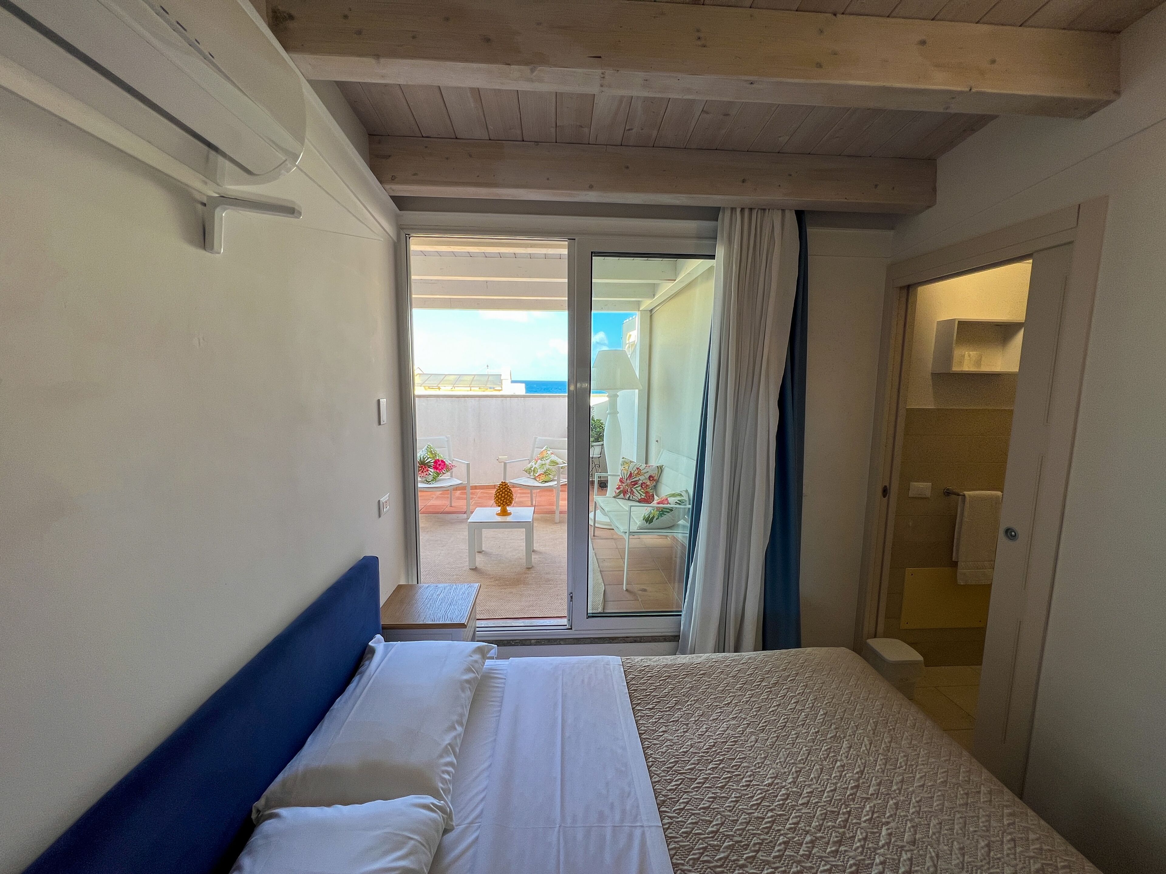 A hotel room with a bed, a small table with a lamp, and a view of an outdoor balcony with chairs and a table.