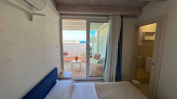 A hotel room with a bed, a small table with a lamp, and a view of an outdoor balcony with chairs and a table.