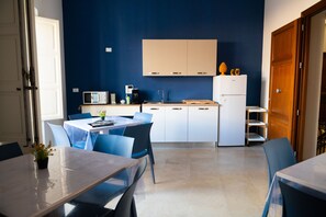 Cocina compacta con armarios blancos, microondas y refrigerador. Cuenta con una zona de comedor con sillas azules y una mesa.