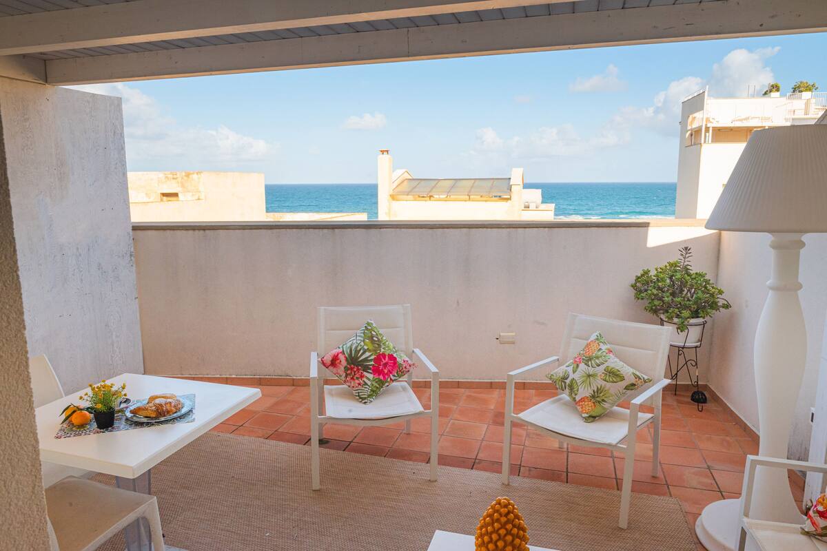 Ein Balkon mit weißen Stühlen, einem Tisch mit Pflanze und Ananas, und Blick aufs Meer.