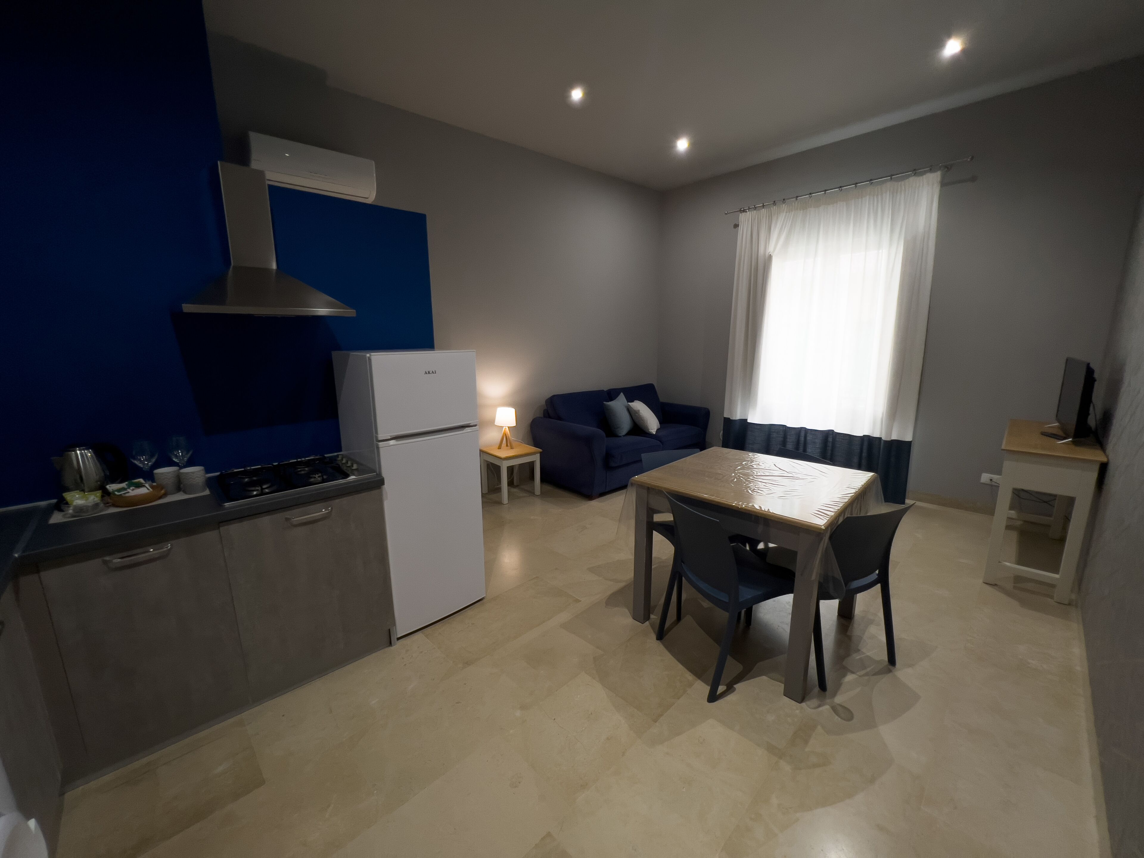 Una cucina compatta con fornello, lavello e frigorifero bianco. È presente un tavolo da pranzo con sedie e un divano blu in un angolo.
