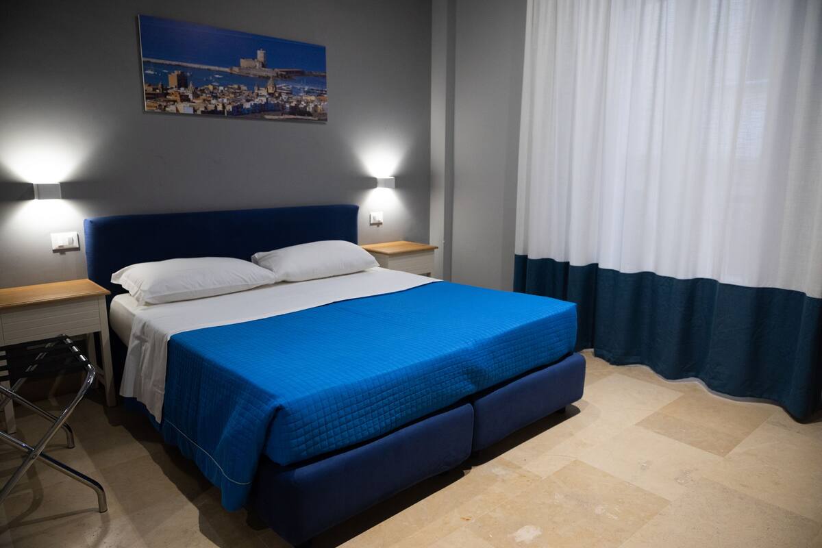Ein Hotelzimmer mit einem großen Bett, zwei Nachttischlampen, einem Nachttisch, einem Sessel und Blick auf eine Stadtlandschaft durch ein mit Vorhängen drapiertes Fenster.
