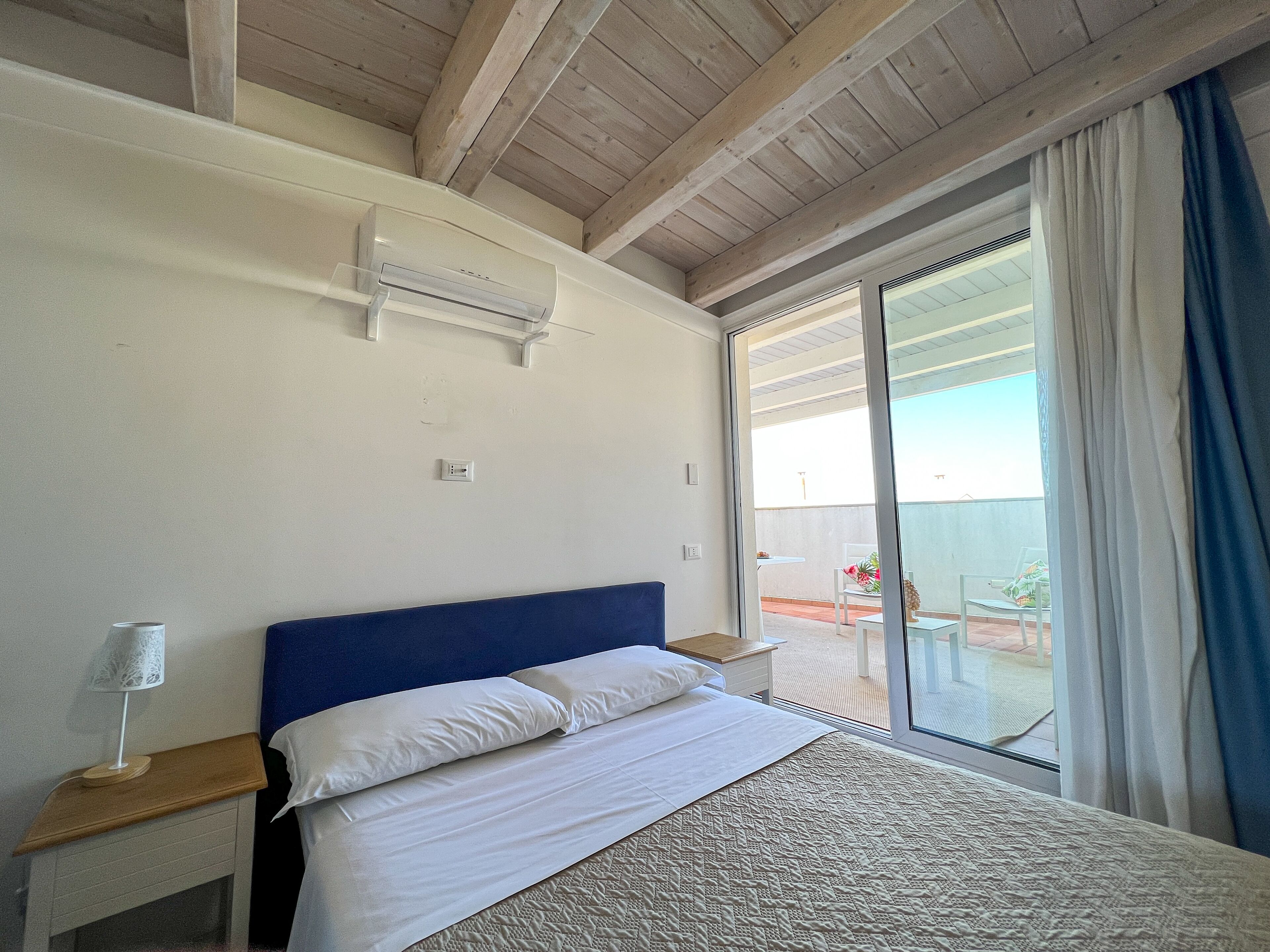 A bedroom with a large bed, a wooden ceiling, a sliding glass door to a balcony, and a small table with a lamp.