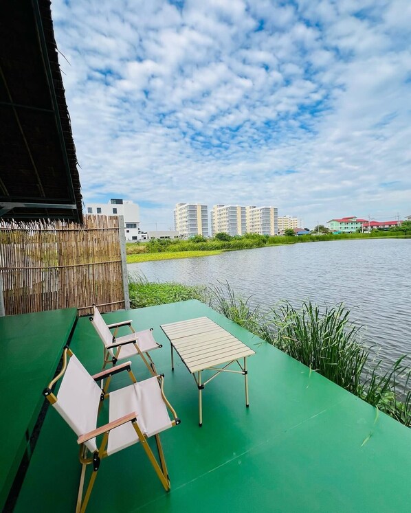 Eine Terrasse mit grünem Boden, zwei Klappstühlen und einem kleinen Tisch. Ein Bambuszaun sowie ein Gewässer mit Gebäuden im Hintergrund sind sichtbar.